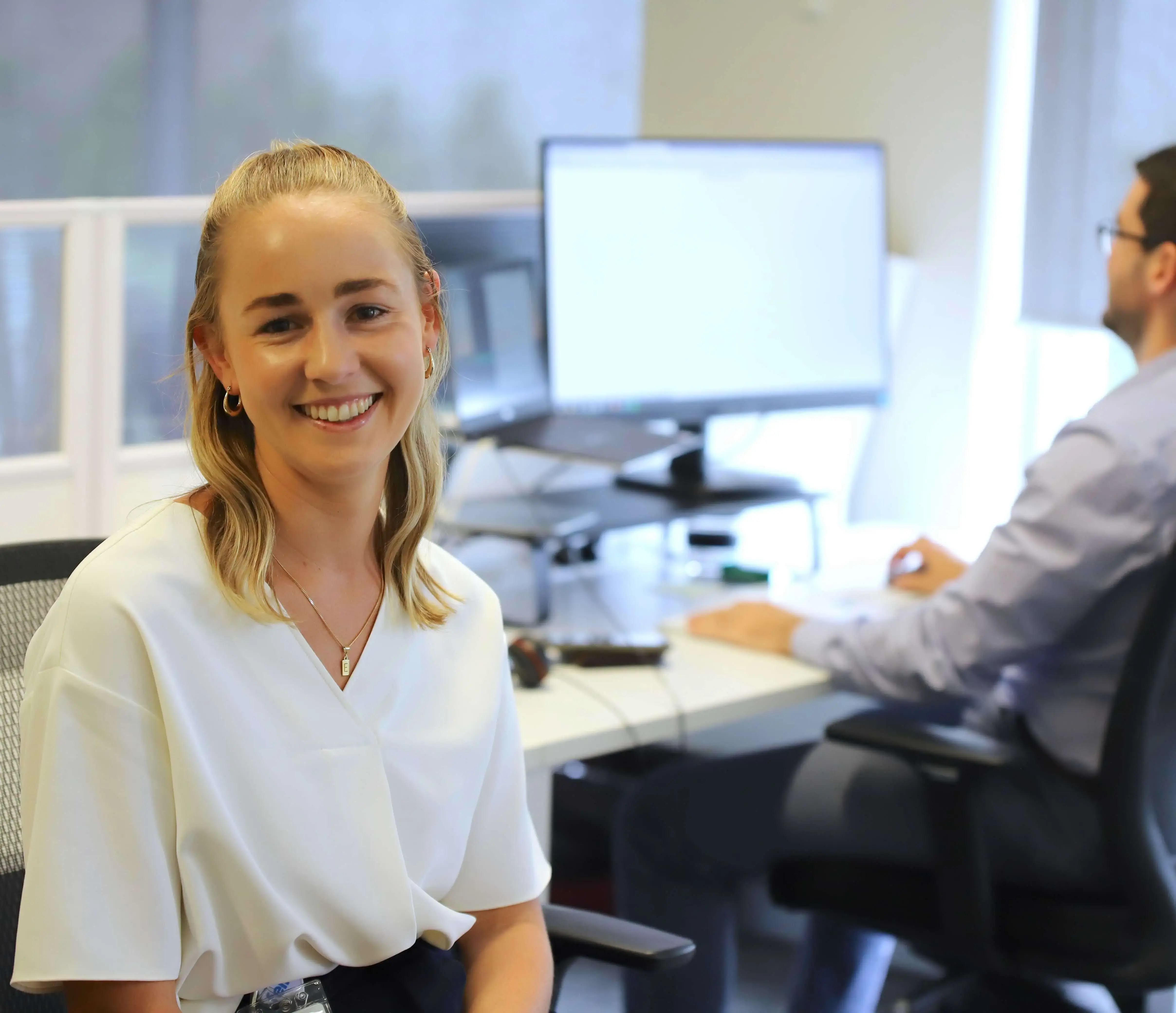 Smiling professional woman in office workspace with colleague at computer in background