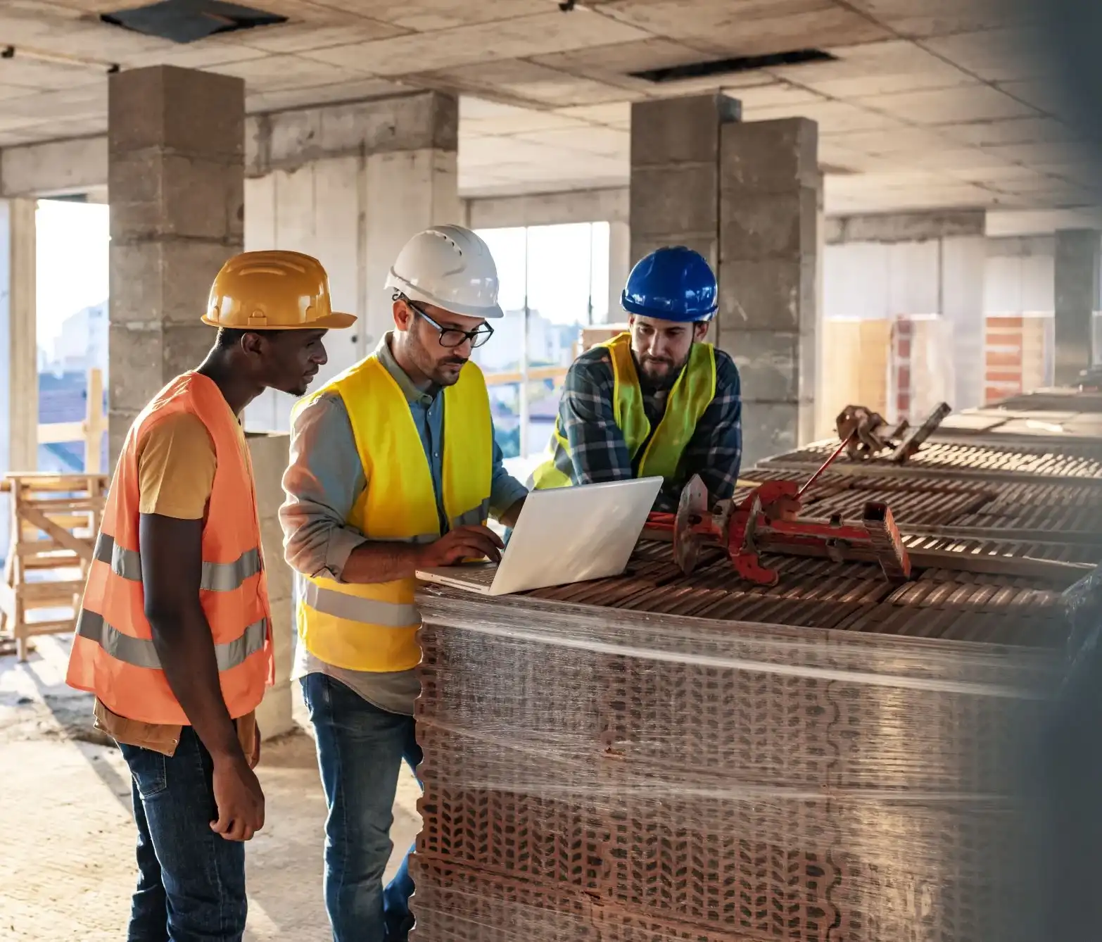 Construction workers wearing safety gear reviewing information on a laptop at an active building site.